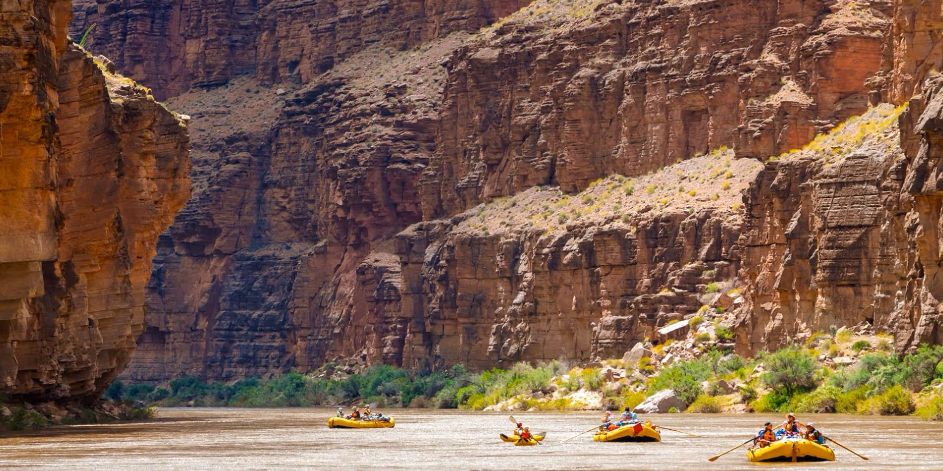 Four yellow rafts with people wearing life jackets rafting on a river surrounded by tall rocky canyon walls.